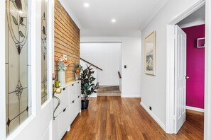 A hallway with a wooden floor, white walls, a brick accent wall, a staircase, and a door leading to a room with a pink wall.