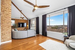 A spacious living room with a large window, a brick wall, a ceiling fan, and a flat-screen TV.