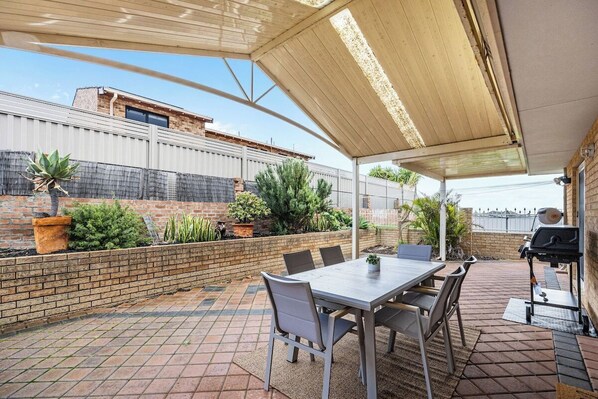 A covered patio with a table and chairs, a grill, and potted plants.