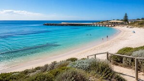 Una spiaggia con acque cristalline turchesi, un molo di legno e una riva sabbiosa con vegetazione rada.