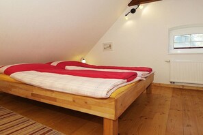 Un simple lit en bois, recouvert d’une couverture rouge, se trouve dans une pièce dont le plafond est en pente et qui possède une fenêtre.