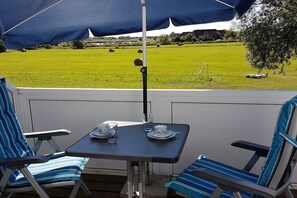 Eine Terrasse mit Tisch und zwei Stühlen, einem blauen Sonnenschirm und Blick auf ein grünes Feld.