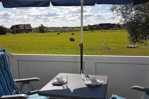 A patio with a table and chairs, a table umbrella, and a view of a green field with hay bales and buildings in the distance. - Holiday apartment with 1 bedroom (Neuharlingersiel)