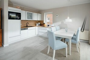 A modern kitchen with white cabinetry, a built-in oven, and a dining area with a white table and blue chairs. - Holiday apartment in Juist with swimming pool (Juist)