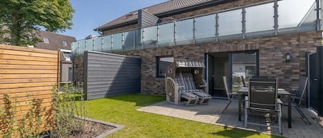 Ein modernes Haus mit Glasbalkon, einer Terrasse und Gartenmöbeln sowie einem gepflegten Rasen.