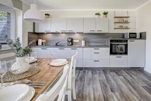 A modern kitchen with white cabinets, a wooden dining table, and a built-in oven. - Ording Seagull Island (St. Peter-Ording)