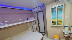 Quarto de hotel com beliche, pequena janela com vista para a paisagem, mesa de cabeceira com abajur e lixeira.