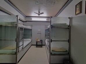 A narrow corridor with bunk beds on either side, a ceiling fan, and a small table with a lamp. - Nice Guest House (Mumbai)