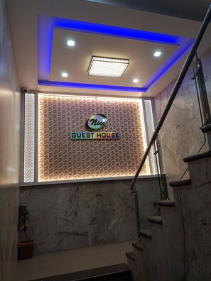 A hotel reception area with a geometric patterned wall, a sign reading 'Nice Guest House,' and a staircase with metal railings.