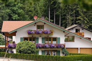 Ein zweistöckiges Chalet mit geneigtem Dach, grünen Fensterläden und Blumenkästen an den Balkonen.