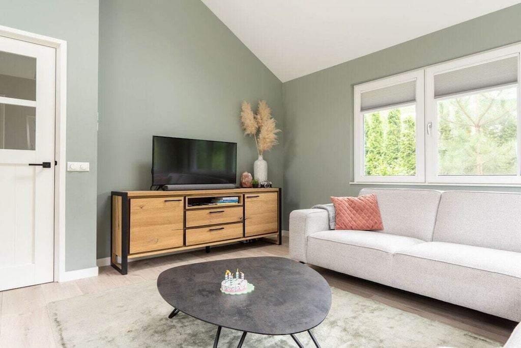 A living room with a flat-screen TV on a wooden cabinet, a grey coffee table with a cake decoration, a white sofa, and a window with a view of trees.