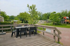 A wooden deck with a table and chairs, surrounded by a wooden railing, overlooking a playground and greenery. - Holiday home with 1 bedroom (Oostkapelle)