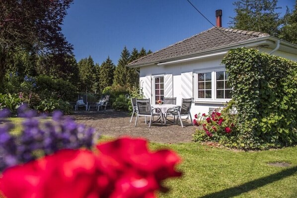 Una casa con fachada blanca, techo de tejas y jardín con flores y arbustos.