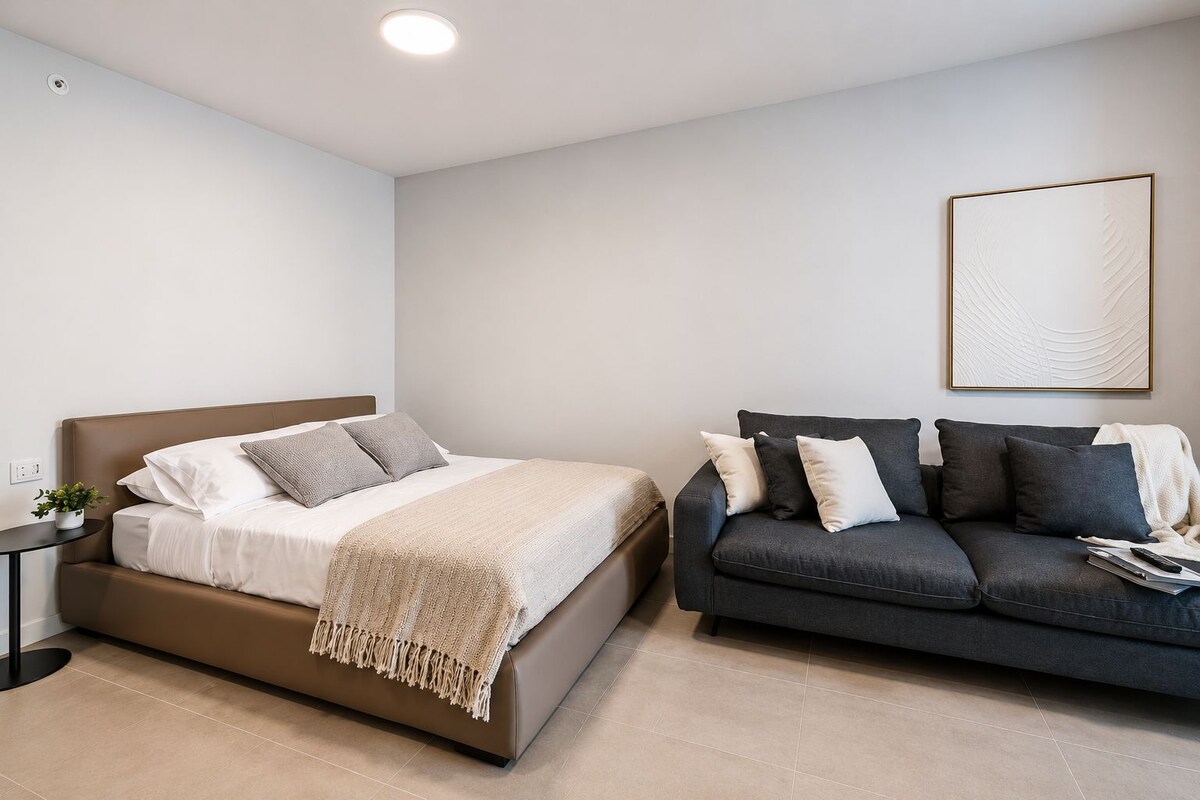 Un dormitorio moderno con cama, un sofá gris oscuro, una mesita con una planta y una obra de arte enmarcada en la pared.