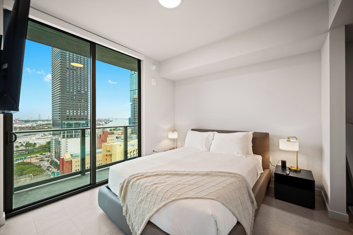 Una habitación de hotel moderna con una cama grande, dos lámparas de noche, vistas de la ciudad a través de ventanales del suelo al techo, y un televisor de pantalla plana montado en la pared.