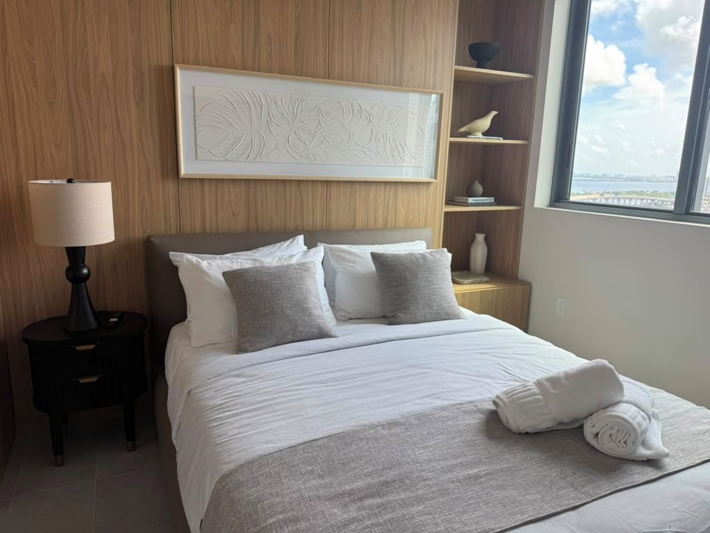A hotel room with a bed, a nightstand, a lamp, a shelf with decorative items, and a window with a view.