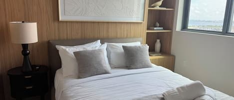 Ett hotellrum med en säng, ett nattduksbord, en lampa, en hylla med dekorativa föremål och ett fönster med utsikt.