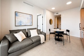 Una sala de estar moderna con un sofá gris, una mesa de comedor con sillas, un cuadro abstracto grande y una cocina con armarios de madera.