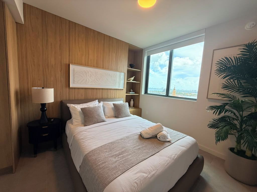 Une chambre d’hôtel avec un grand lit, une table de chevet, une lampe, une bibliothèque, une grande fenêtre donnant sur la ville et une plante en pot.
