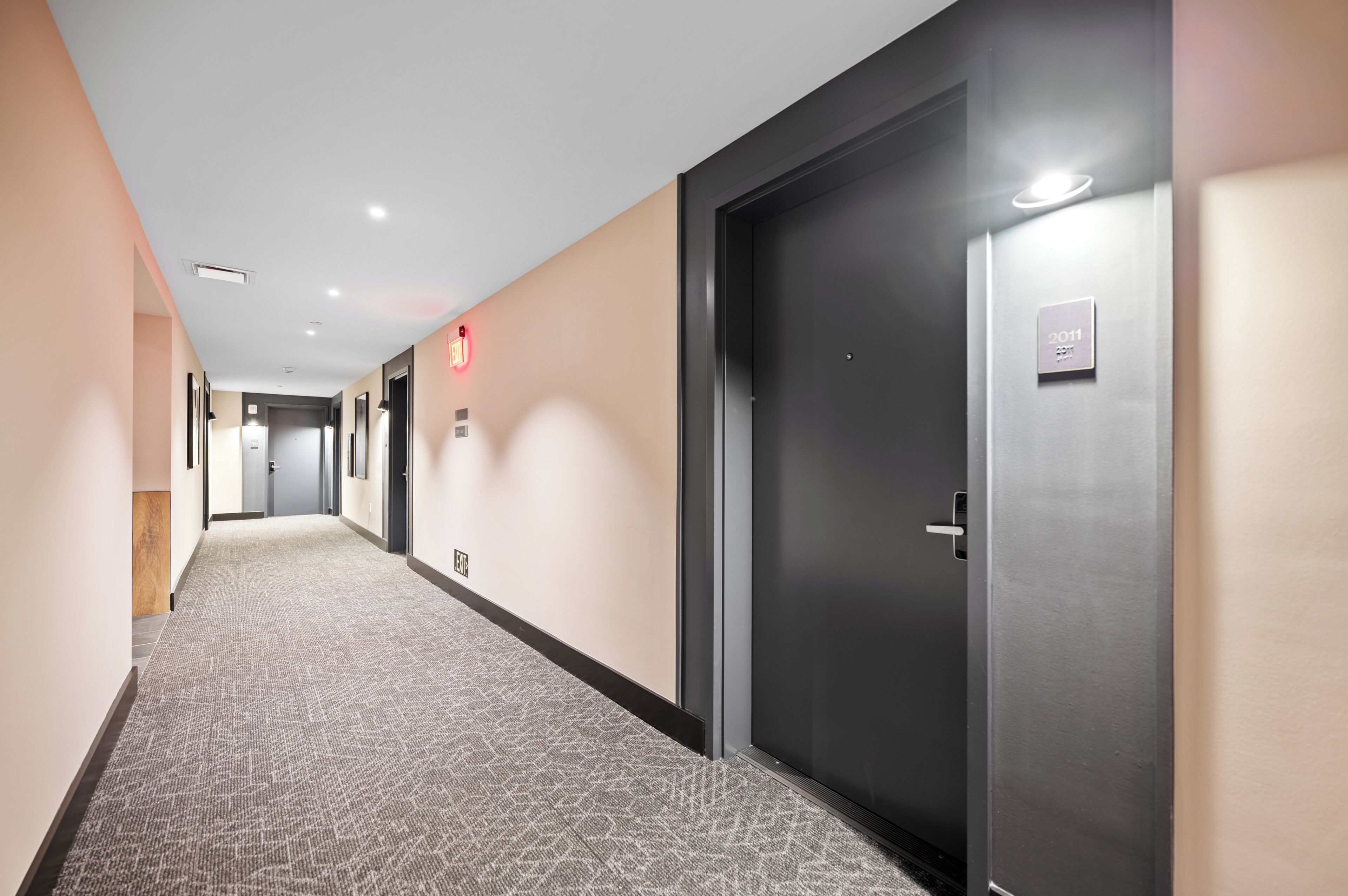Un pasillo de hotel con pisos alfombrados, una puerta oscura a la derecha y una señal de salida en la pared.