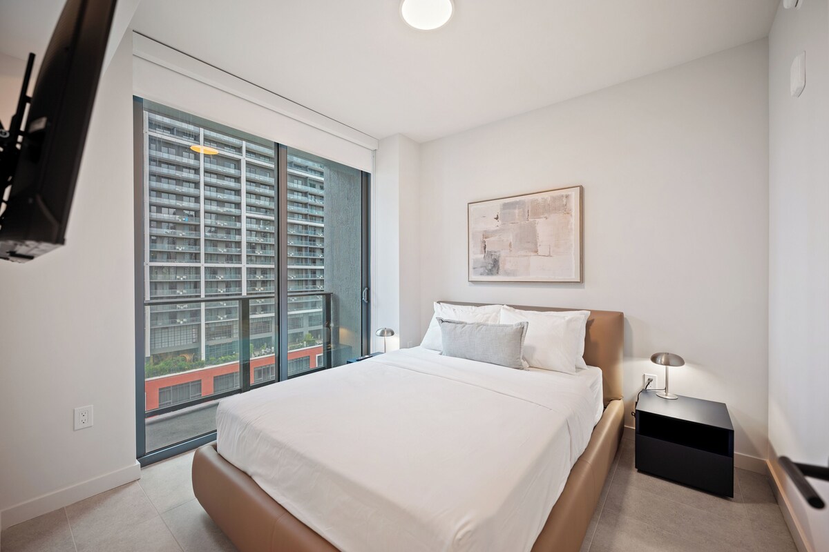 Habitación de hotel moderna con una cama grande, un televisor de pantalla plana montado en la pared, una ventana con vistas a un edificio alto y una obra de arte minimalista en la pared.