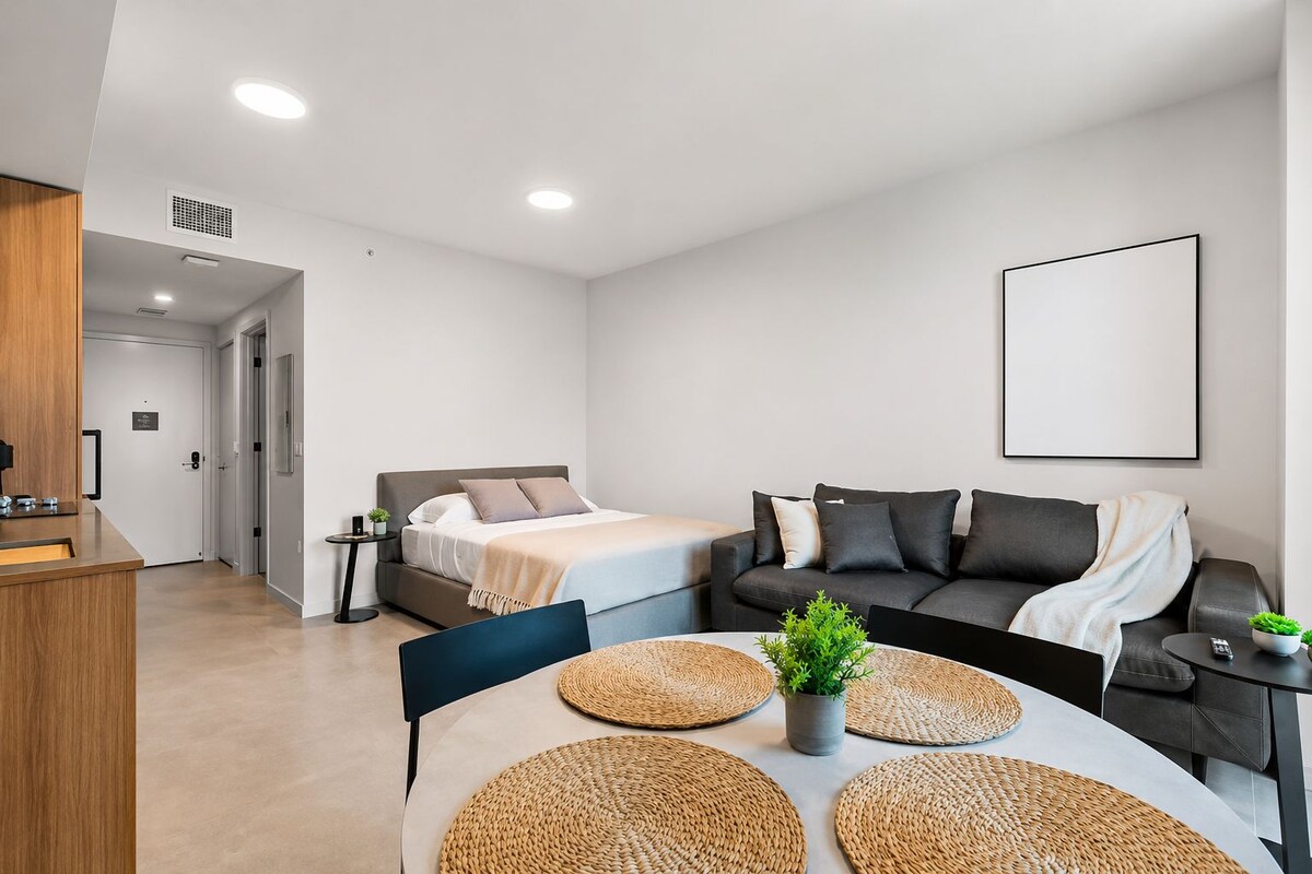 Una habitación de hotel moderna con cama, sofá, mesa de comedor con individuales y una pequeña planta en maceta.