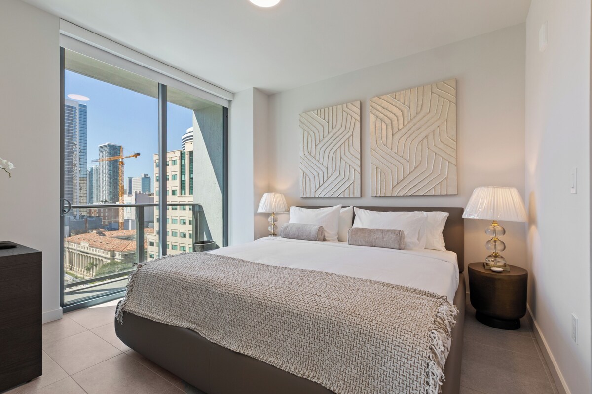 Una habitación de hotel moderna con una cama grande, dos lámparas de noche, vistas del paisaje urbano a través de ventanales del suelo al techo y arte decorativo en la pared.