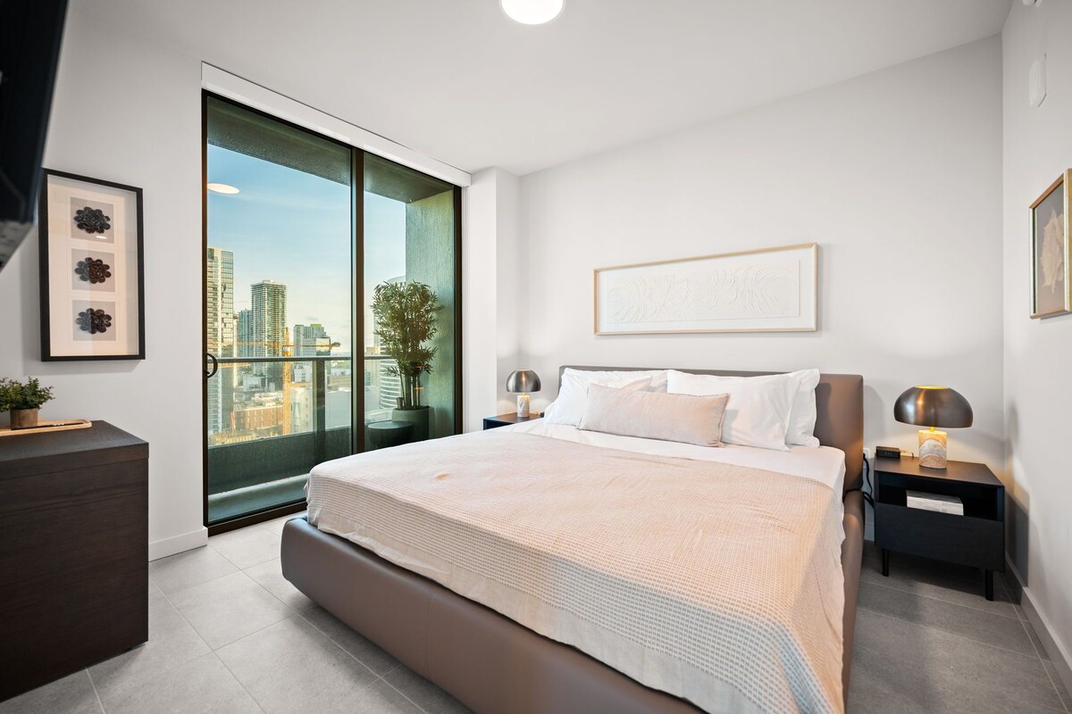 Un dormitorio moderno con una cama grande, vistas a la ciudad, una planta en maceta y una mesita de noche con una lámpara.