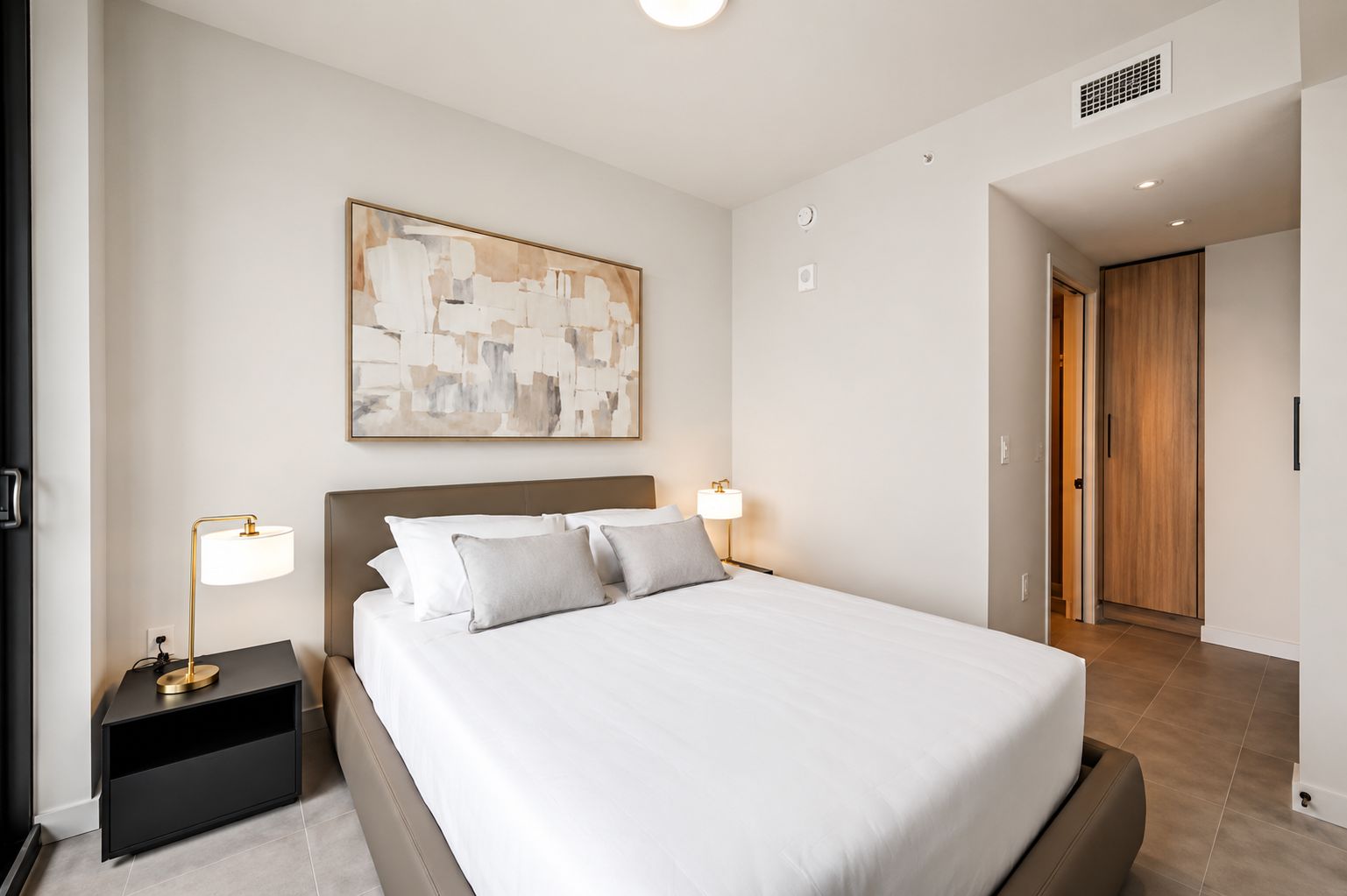 A modern hotel room with a large bed, bedside lamps, a painting on the wall, and a wooden door.