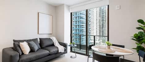 Uma sala de estar moderna com um sofá cinzento, uma mesa de jantar com cadeiras e uma grande janela com vista para a cidade.