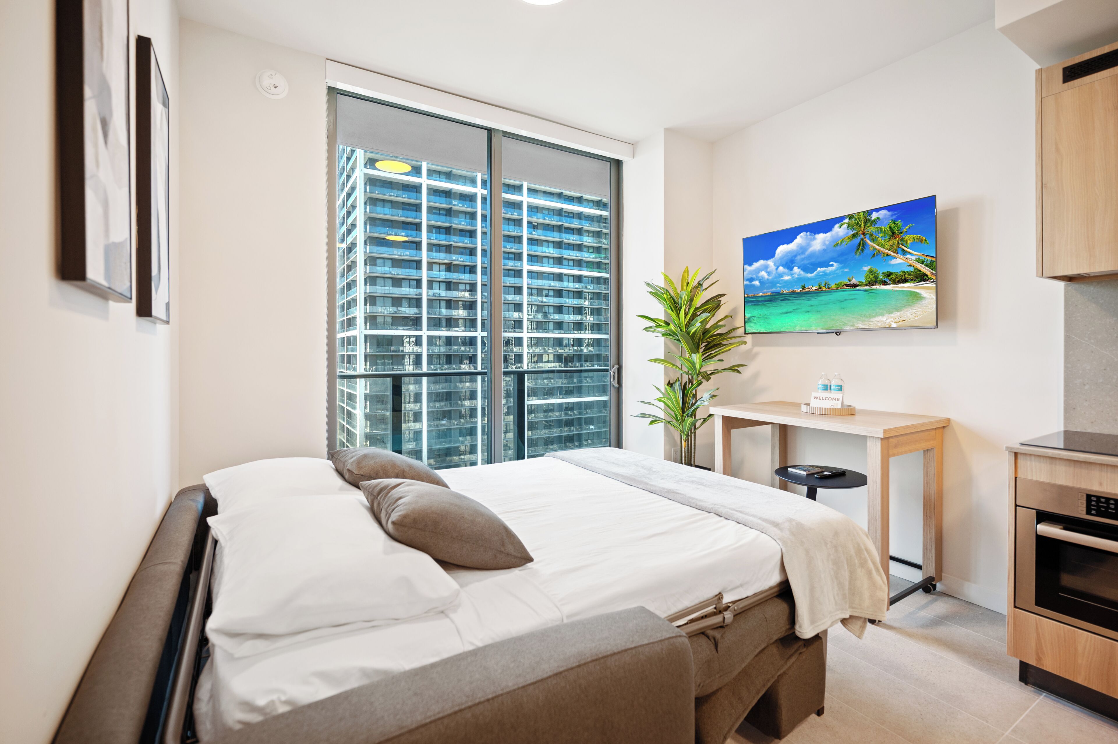 A modern hotel room with a large bed, a desk, a television, and a view of a high-rise building.