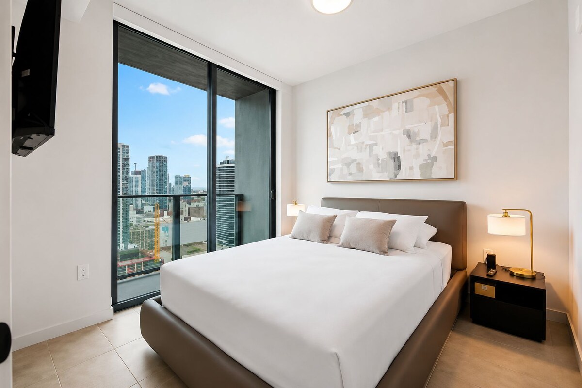 Habitación de hotel moderna con una cama grande, vista a la ciudad, un cuadro en la pared y una mesita de noche con una lámpara.