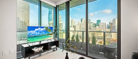 A modern living room with a large TV, a bookshelf, and a view of a cityscape through floor-to-ceiling windows.