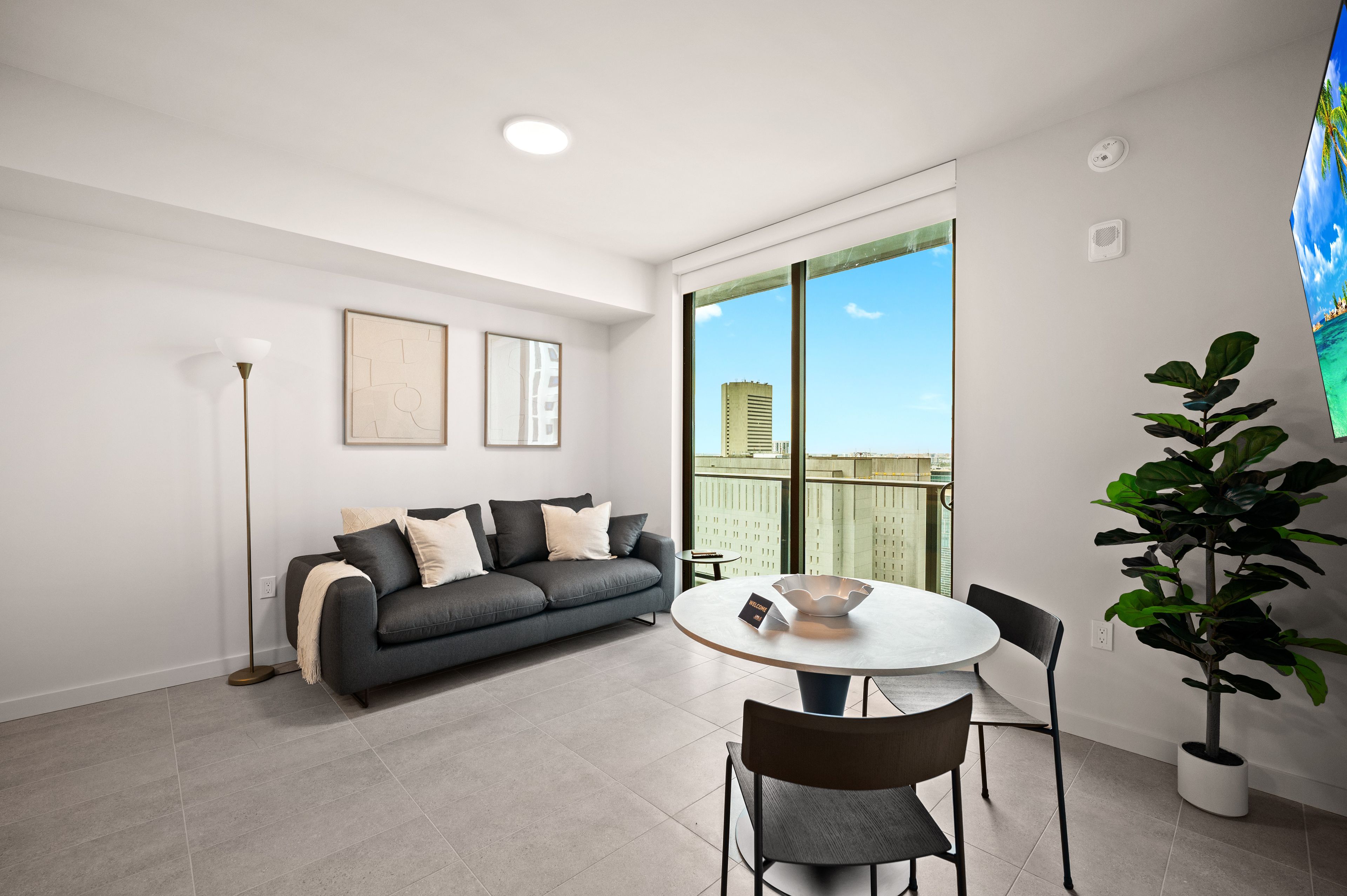 A modern living room with a grey sofa, a round table, and chairs, a floor lamp, and a large window with a city view.