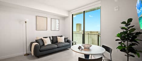 A modern living room with a grey sofa, a round table, and chairs, a floor lamp, and a large window with a city view.