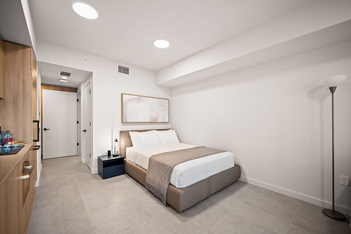 Habitación de hotel moderna con cama, mesita de noche, lámpara y vista al pasillo.