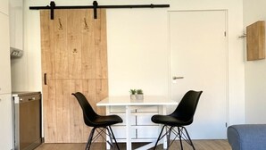 Cozinha compacta com mesa branca e duas cadeiras pretas, porta deslizante de madeira e um sofá azul na área de estar adjacente.