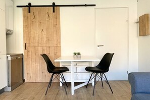 Eine kleine Küche mit einem weißen Tisch und zwei schwarzen Stühlen, einer Holztür mit Schiebevorrichtung und einem blauen Sofa im angrenzenden Wohnbereich.