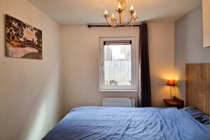 Une chambre à coucher avec un lit, une fenêtre, un radiateur, une table de chevet et une lampe.