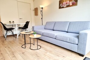 Un salon avec un canapé gris, deux fauteuils noirs, une petite table ronde ornée d’une plante, une table blanche et un lampadaire.