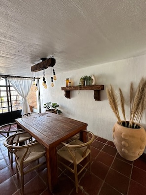 A rustic dining area with a wooden table and wicker chairs, a hanging light fixture, and a large potted plant.