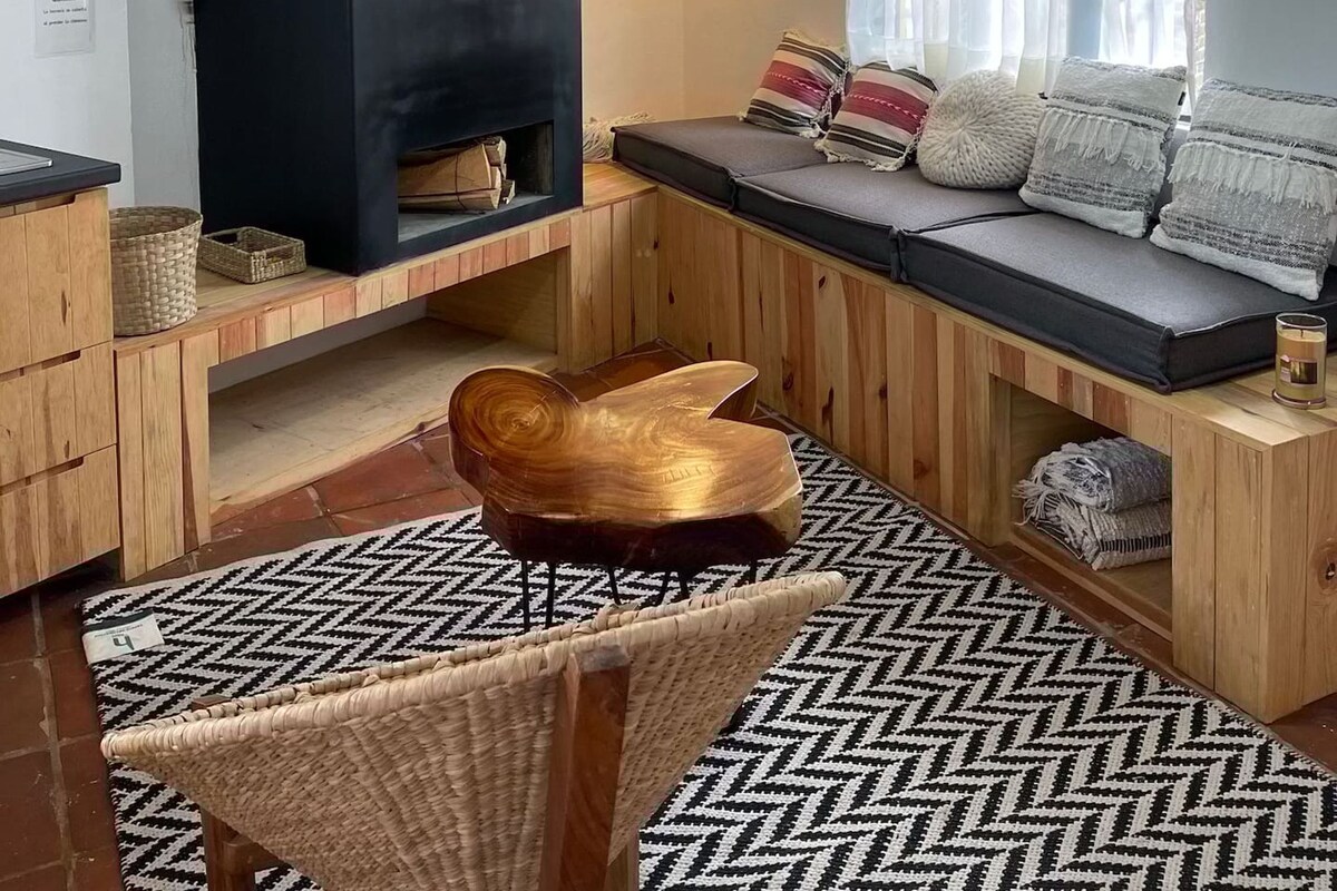 A cozy living room with a black fireplace, a woven chair, a patterned rug, and a cushioned bench with pillows.