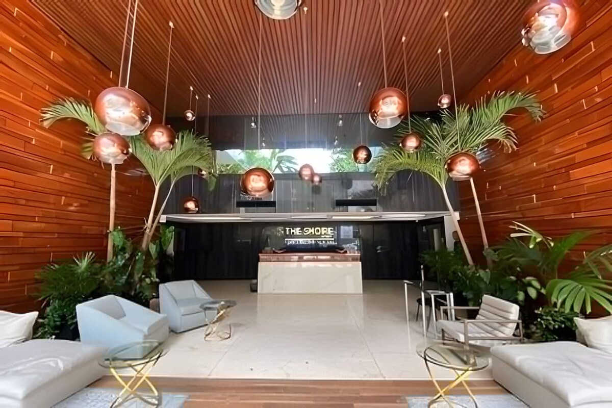 Ein modernes Interieur mit einer Rezeption mit der Aufschrift 'THE SHORE', einer Sitzecke mit Sofas und Sesseln sowie dekorativer Beleuchtung.