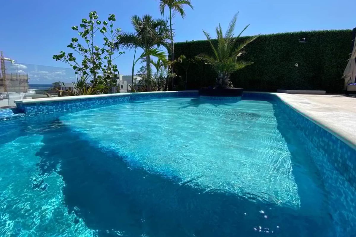 Ein klares, blaues Schwimmbecken mit einer gefliesten Kante, umgeben von Grünpflanzen und einer Hecke.