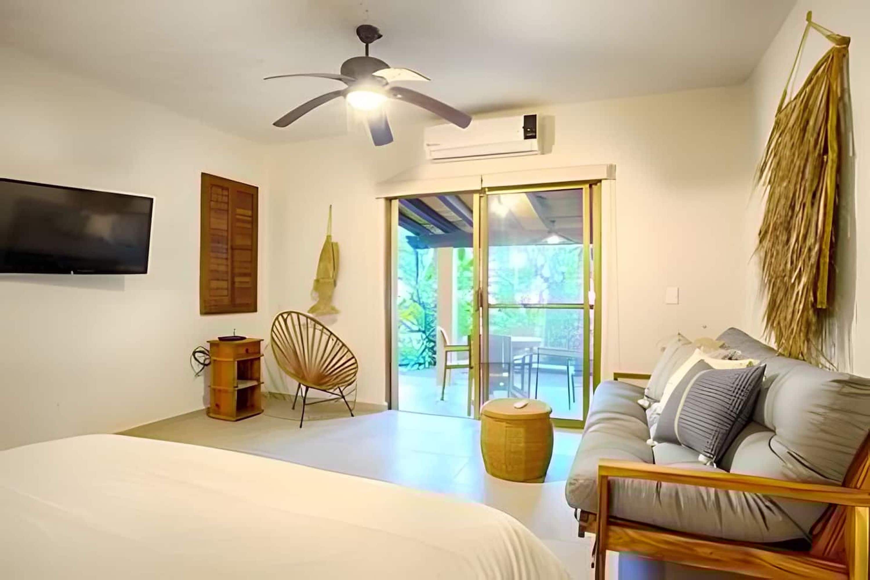 A bedroom with a bed, a ceiling fan, a TV mounted on the wall, a wooden chair, a sofa, and a sliding glass door leading to an outdoor area.