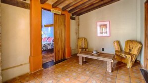 Une pièce comprenant un banc en bois, deux chaises, un lit avec une literie à motifs et une porte donnant sur une autre pièce.