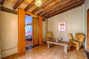 A room with a wooden bench, two chairs, a bed with patterned bedding, and a door leading to another room.