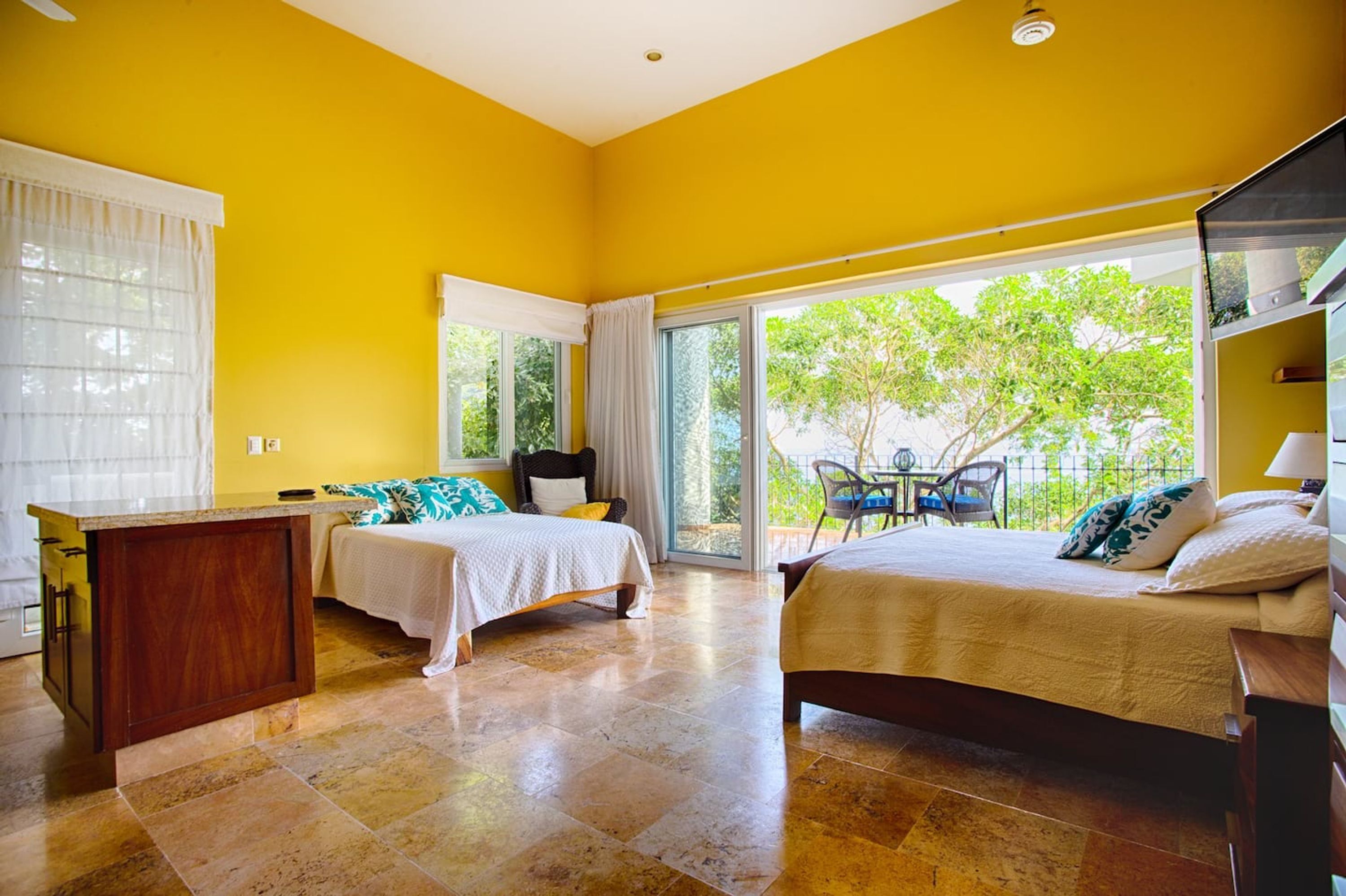 A hotel room with two beds, a dining area, and a view of greenery outside.