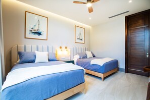 Een hotelkamer met twee bedden, een plafondventilator en twee ingelijste foto’s van zeilboten aan de muur.
