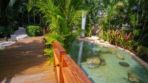 A wooden deck leading to a clear pool surrounded by lush greenery.